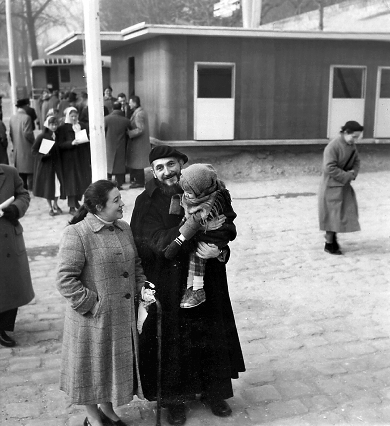 L’abbé Pierre devant la maison démontable Les Jours Meilleurs en compagnie de la famille à laquelle elle est destinée, quai Alexandre-III, Paris, février 1956.