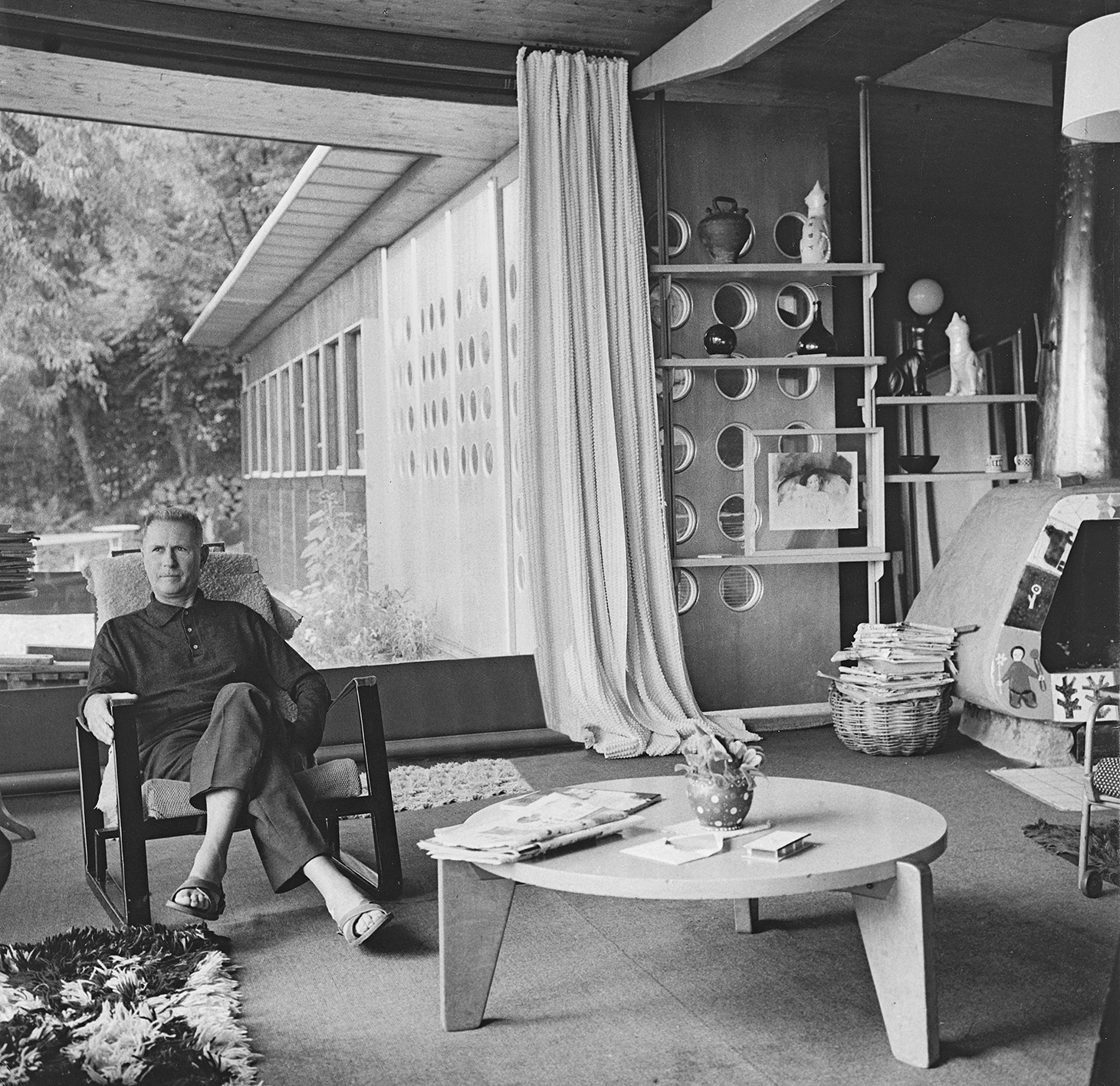 Jean Prouvé dans le coin-séjour de la maison, espace équipé d’une étagère équilibrée à crémaillères, d’un fauteuil Cité et d’un guéridon bas GB 21 de Jean Prouvé, Le Haut-du-Lièvre, Nancy, 1962.
